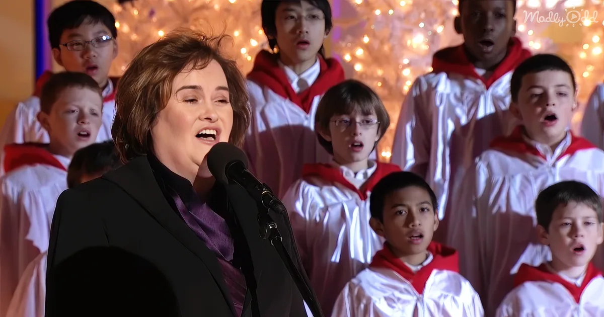 A woman sings into a microphone, surrounded by children in choir robes against a festive backdrop.