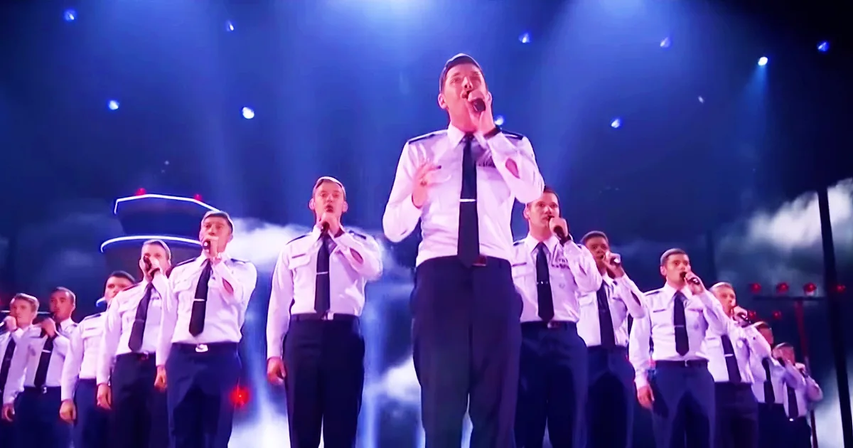 A man in a white shirt and black tie sings into a microphone, surrounded by a choir in uniform.