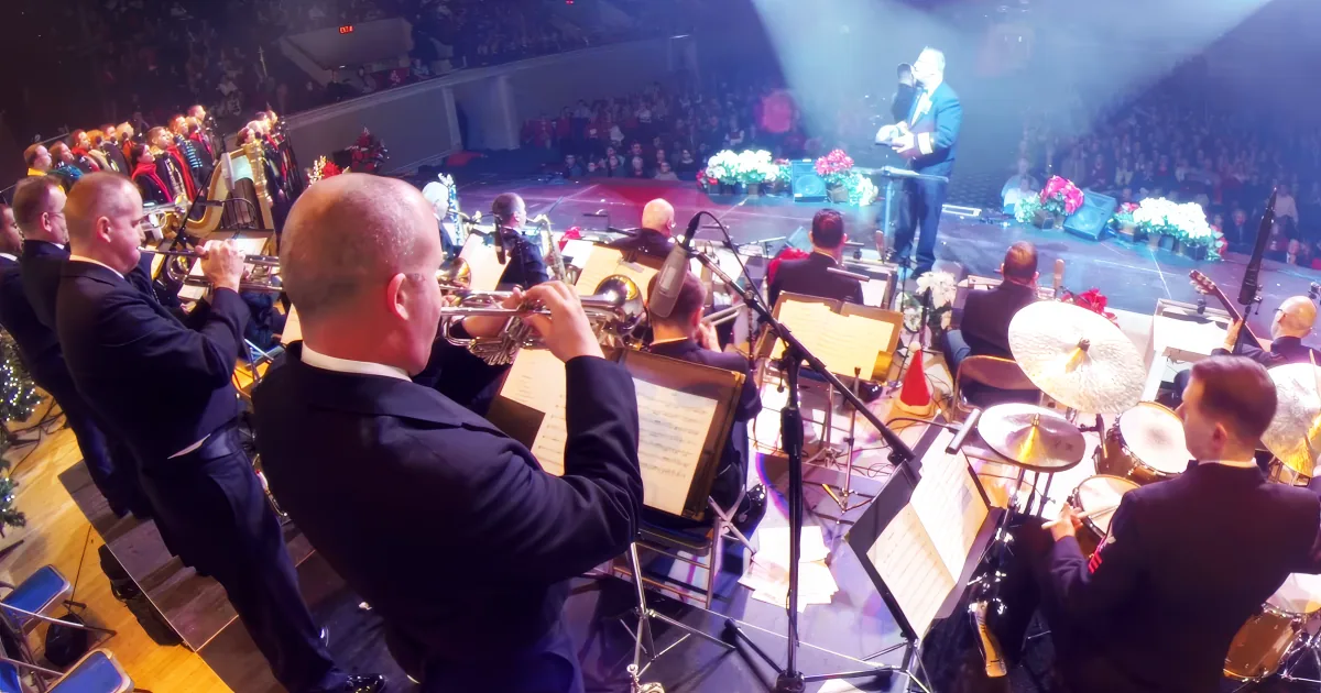 A conductor leads an orchestra on stage with musicians playing instruments and sheet music stands.