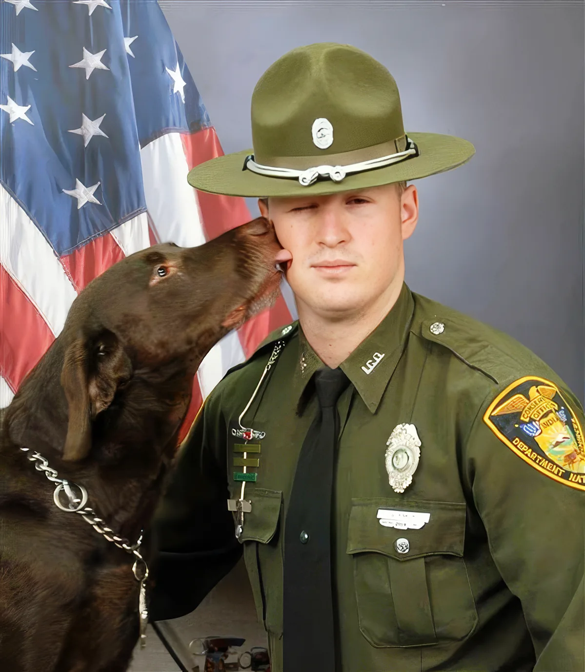 A uniformed officer in a green uniform is being kissed on the cheek by a chocolate Labrador retriever.
