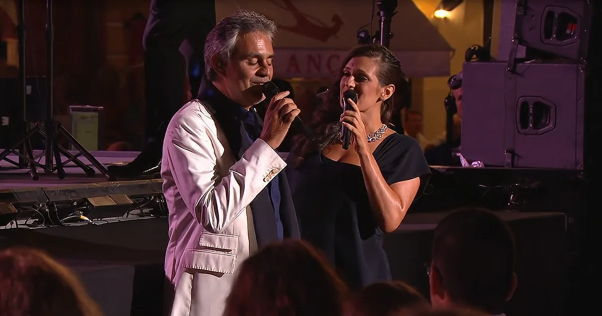 A man and woman sing on stage, both holding microphones, with an audience in the foreground and stage equipment in the...