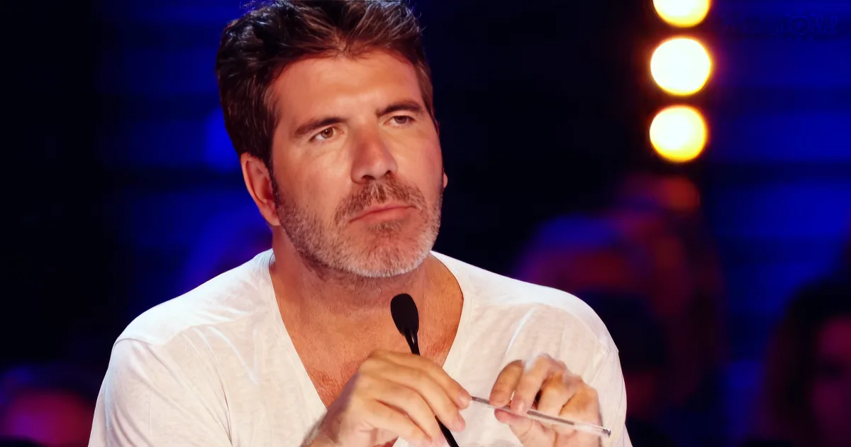 A man with a beard and white shirt sits at a table with a microphone, appearing to listen intently.