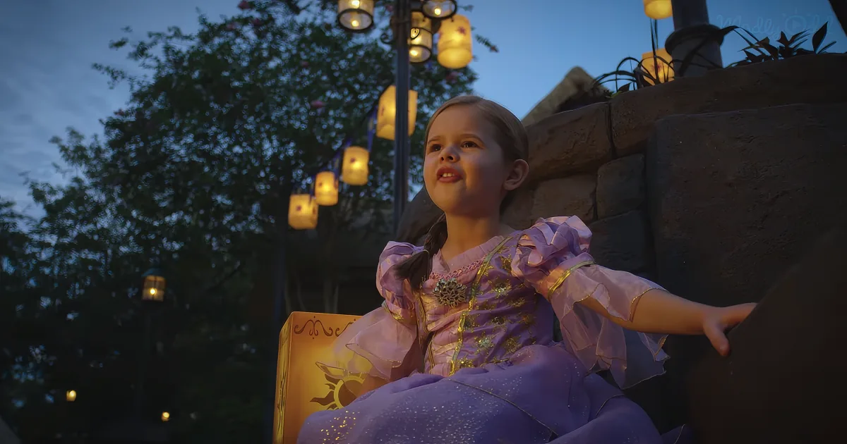 A child in a purple princess dress sits on a stone wall, surrounded by lanterns, under a twilight sky.