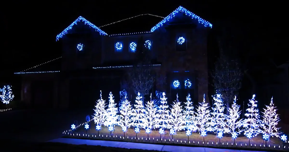 A house adorned with blue Christmas lights and illuminated trees creates a festive winter scene.