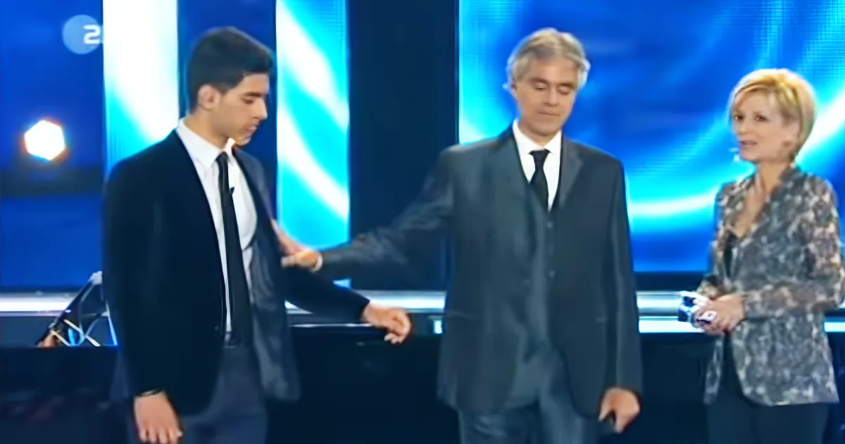 A man in a suit is being patted on the back by another man in a suit, with a woman in a patterned blazer standing nearby.