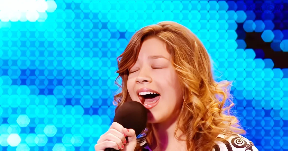 A person with curly hair singing into a microphone against a blue, hexagonal-patterned background.