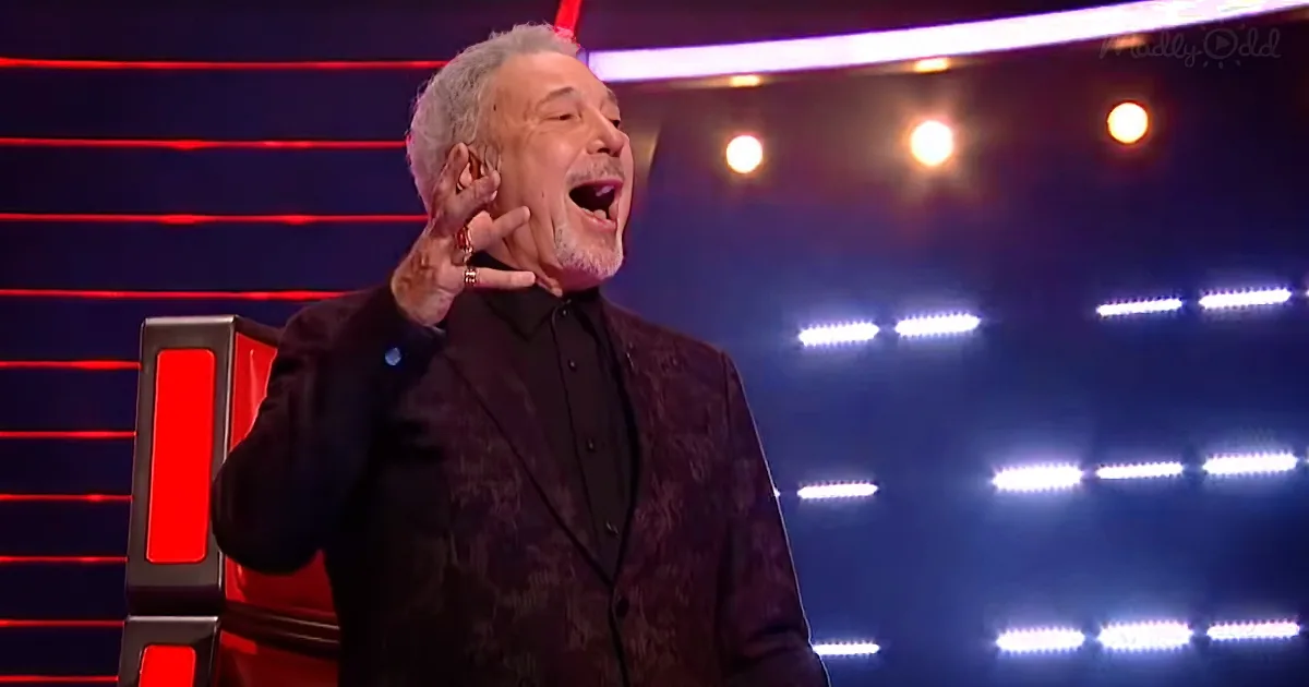 A man in a black suit is singing passionately on a stage with red and blue lighting.