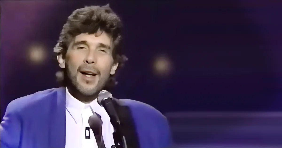 A man in a blue suit sings into a microphone on stage with a dark background.
