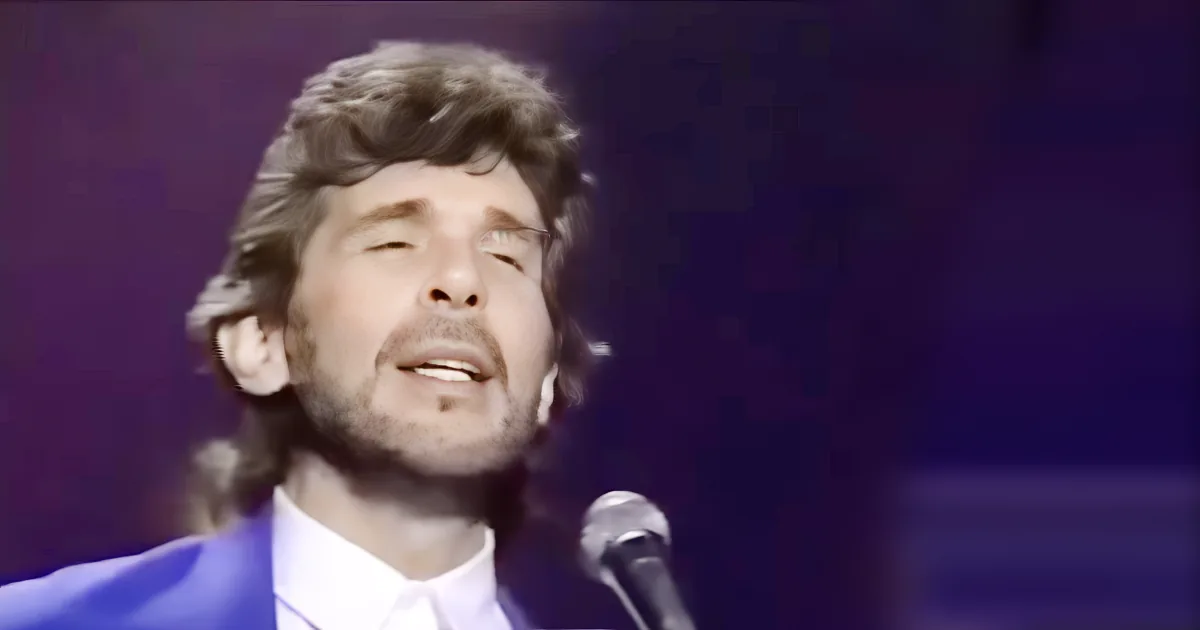 A man with curly hair and a blue suit sings into a microphone against a dark background.