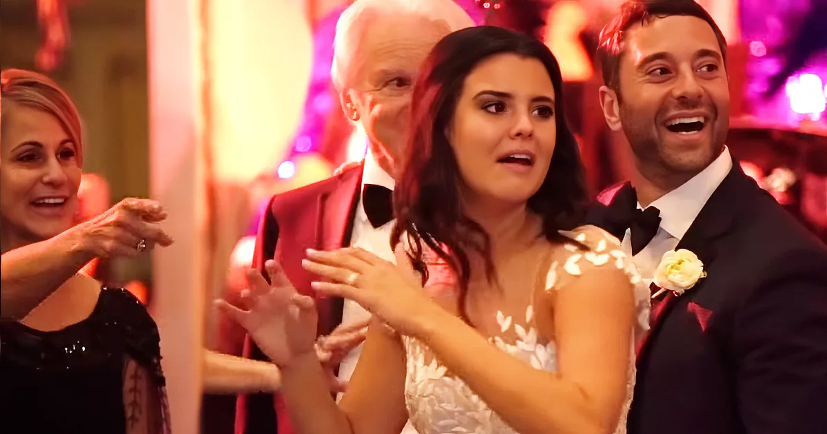 The bride, in a white dress, is clapping with her hands raised, surrounded by guests in formal attire.