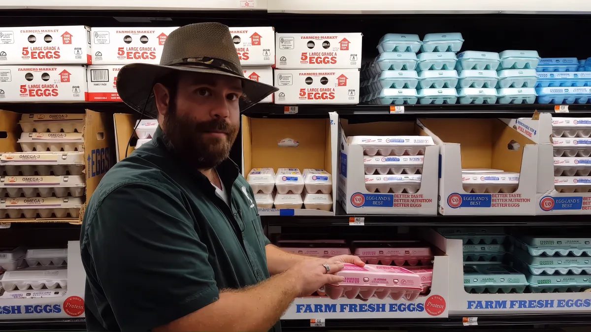 A man in a hat holds a pink egg carton in a grocery store egg aisle.