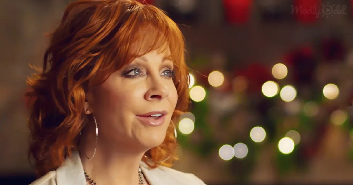 A woman with red hair and hoop earrings smiles softly, with blurred lights and decorations in the background.