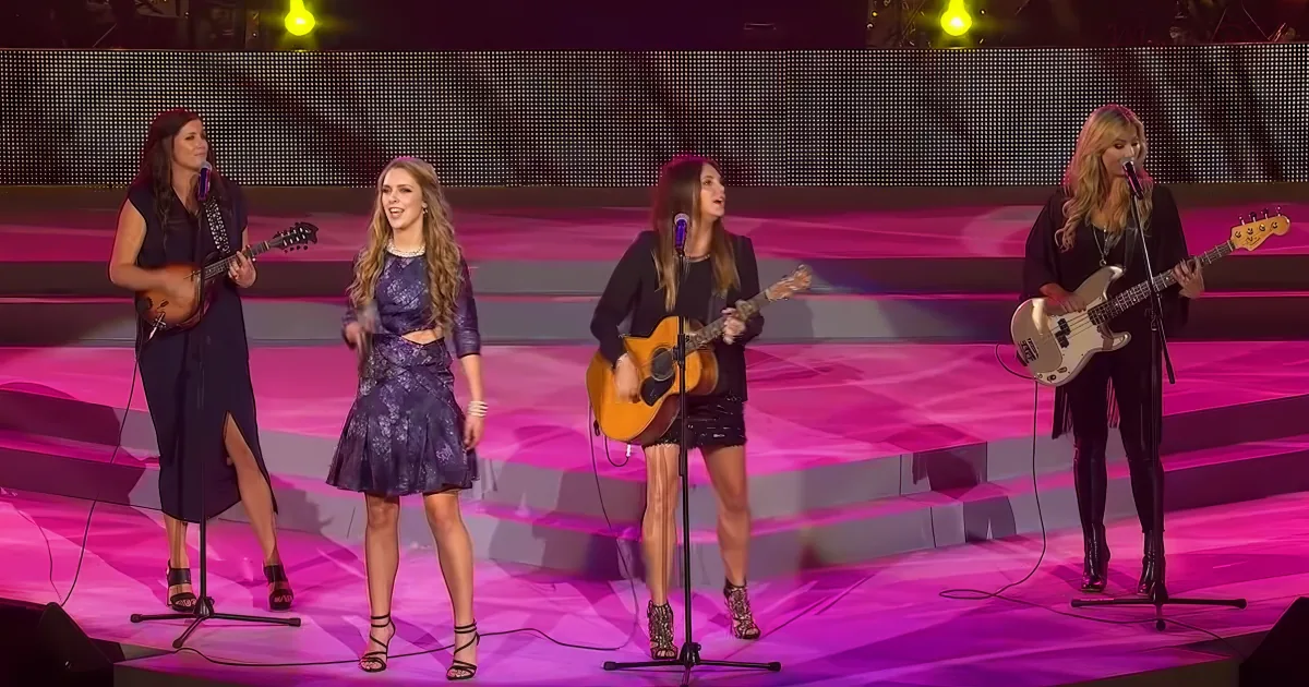 Four women perform on stage with microphones, guitars, and a bass, under vibrant pink lighting.