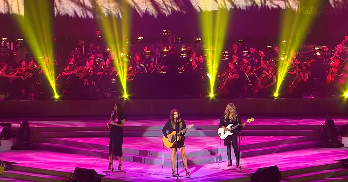 Three women perform on stage with guitars, illuminated by vibrant pink and yellow lights.