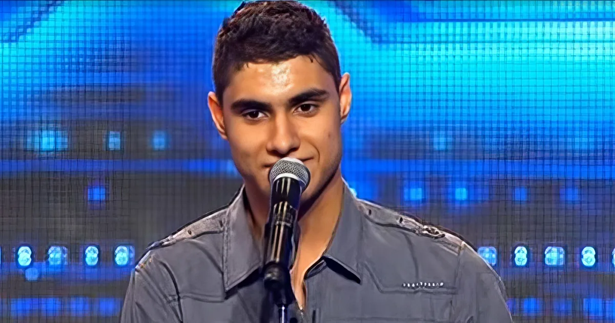 A young man in a gray shirt sings into a microphone against a blue-lit background.