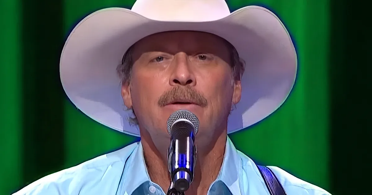 A man in a white cowboy hat and light blue shirt sings into a microphone against a green backdrop.