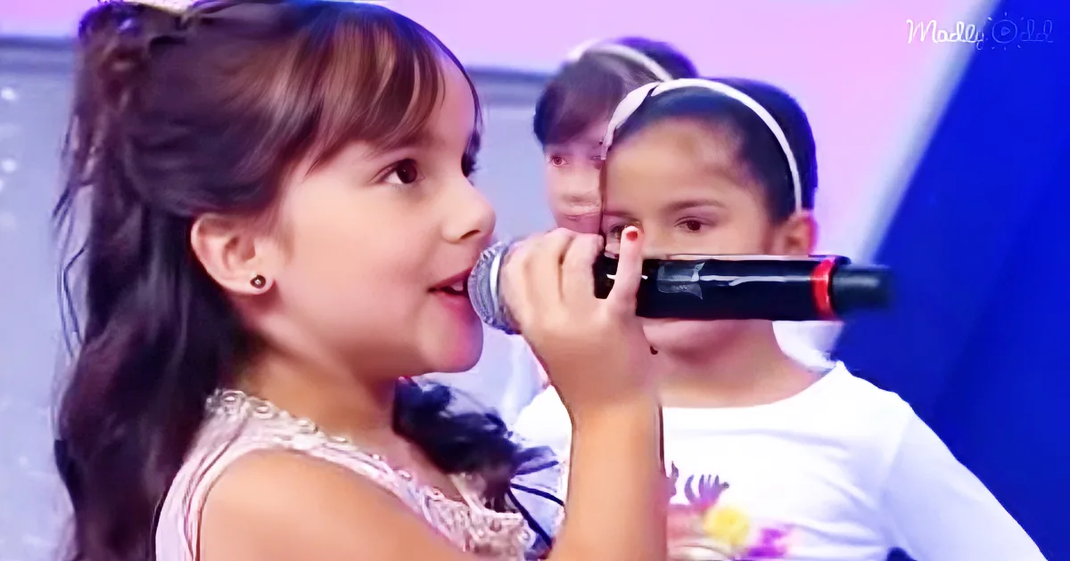 A young girl sings into a microphone, wearing a white dress, with another child in the background.