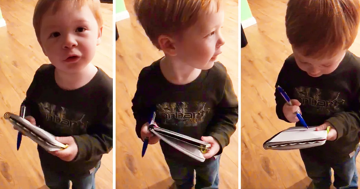 A young boy in a black shirt holds a pen and notebook, looking down and to the side.