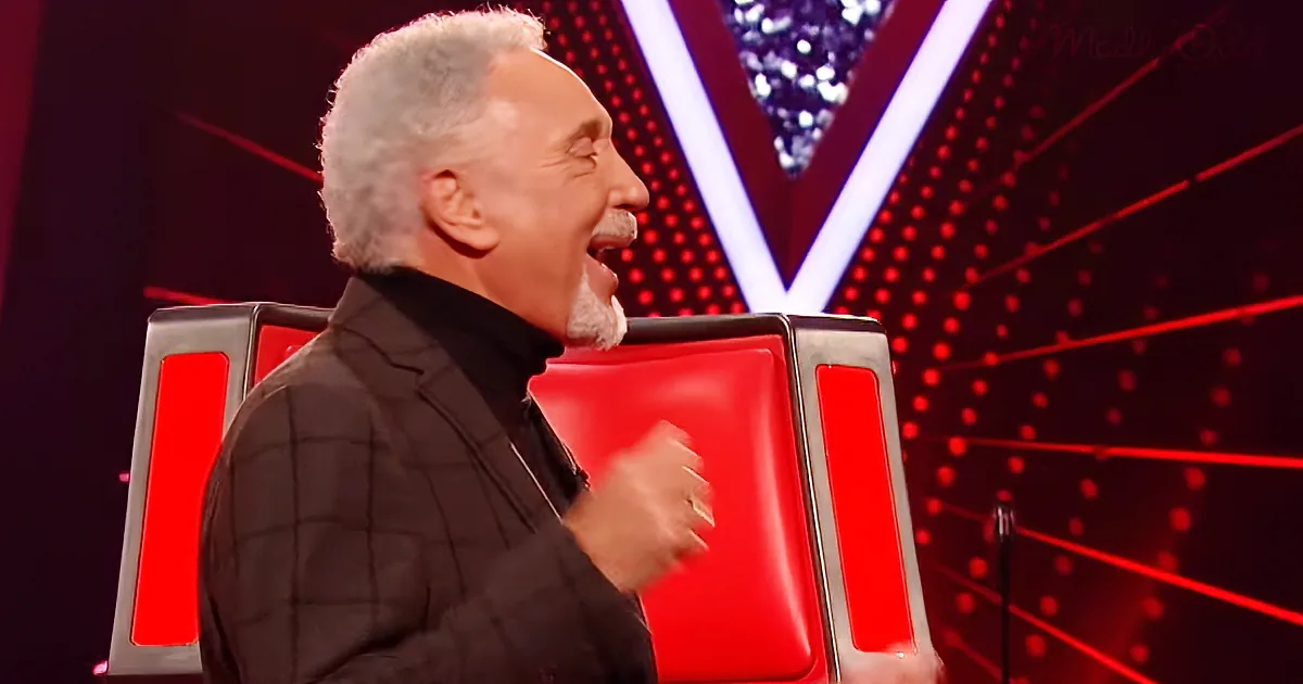 A man with white hair and a beard is smiling and gesturing with his hand while sitting in a red chair on a stage.