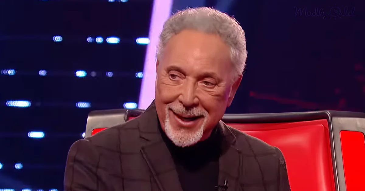 A smiling man with gray hair and a beard sits in a red chair, wearing a black jacket and black turtleneck.