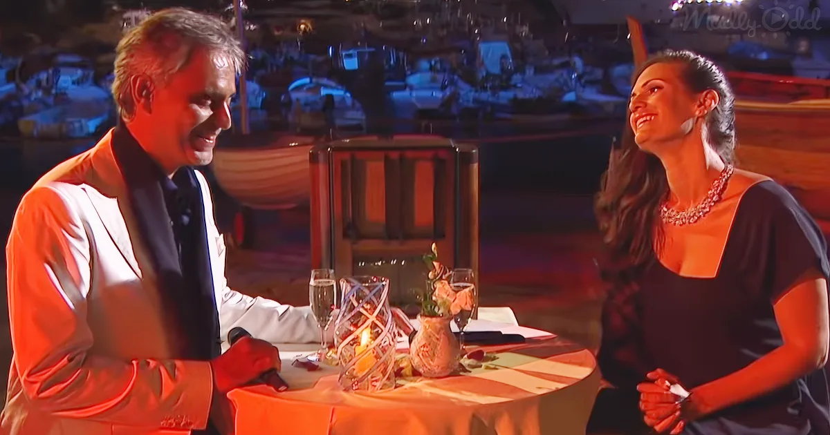 A man in a white suit and a woman in a black dress are seated at a table with a romantic setup, smiling at each other.