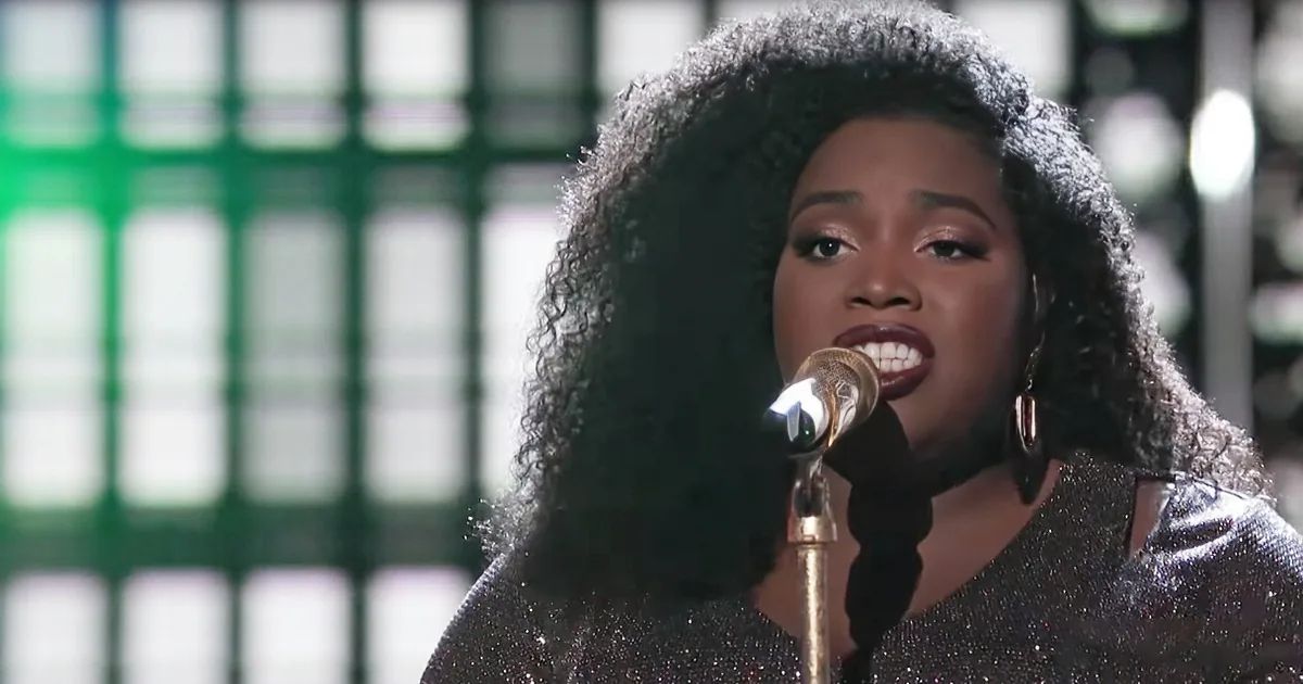 A person with curly hair wearing a sparkly outfit sings into a microphone against a green grid background.