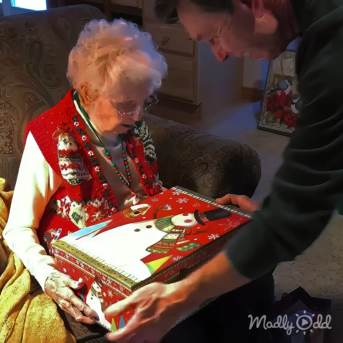 An elderly woman in a red sweater is opening a Christmas-themed gift box with assistance.