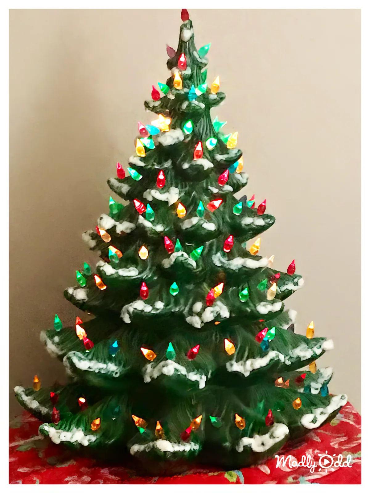 A ceramic Christmas tree adorned with colorful lights and snow-like decorations stands against a plain wall.