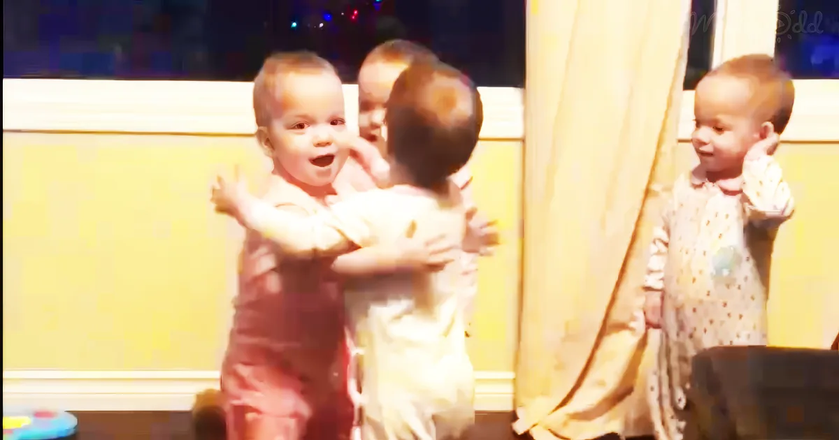A baby in red pajamas is being hugged by another baby in white pajamas, while a third baby in white pajamas looks on.