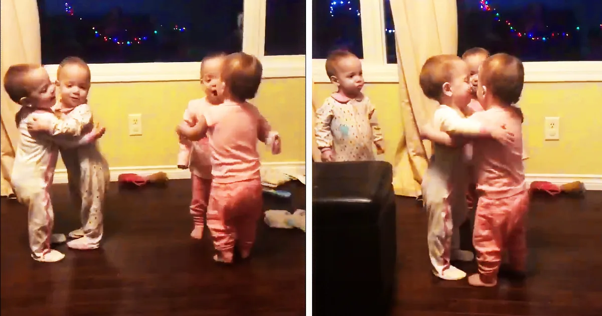 Two babies in pajamas are hugging and dancing together in a room with a window and Christmas lights.