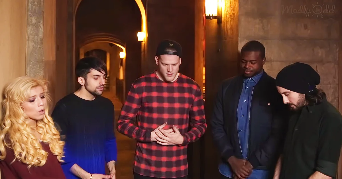 A man in a red plaid shirt stands with hands clasped, surrounded by four others in a dimly lit hallway.