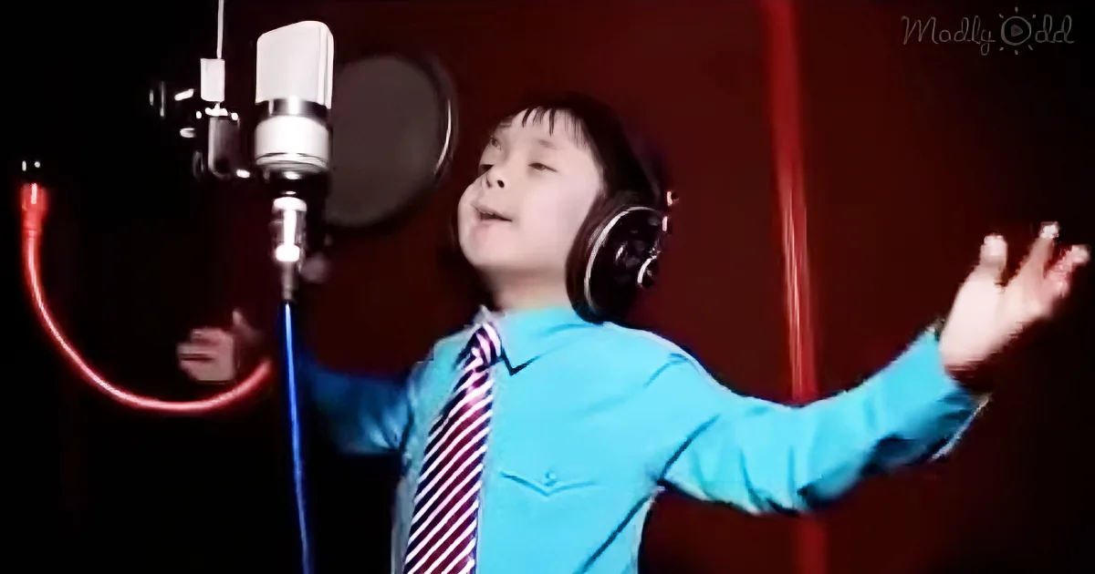 A child in a blue shirt and tie sings into a microphone, wearing headphones, with arms outstretched.