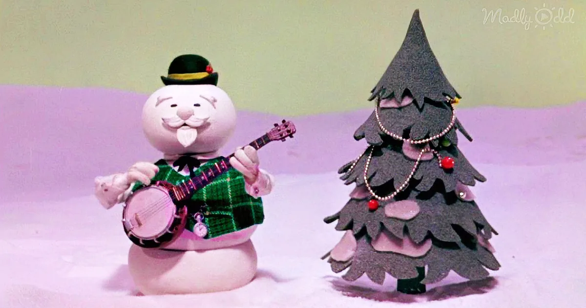 A snowman in a green plaid shirt plays a banjo next to a decorated Christmas tree.