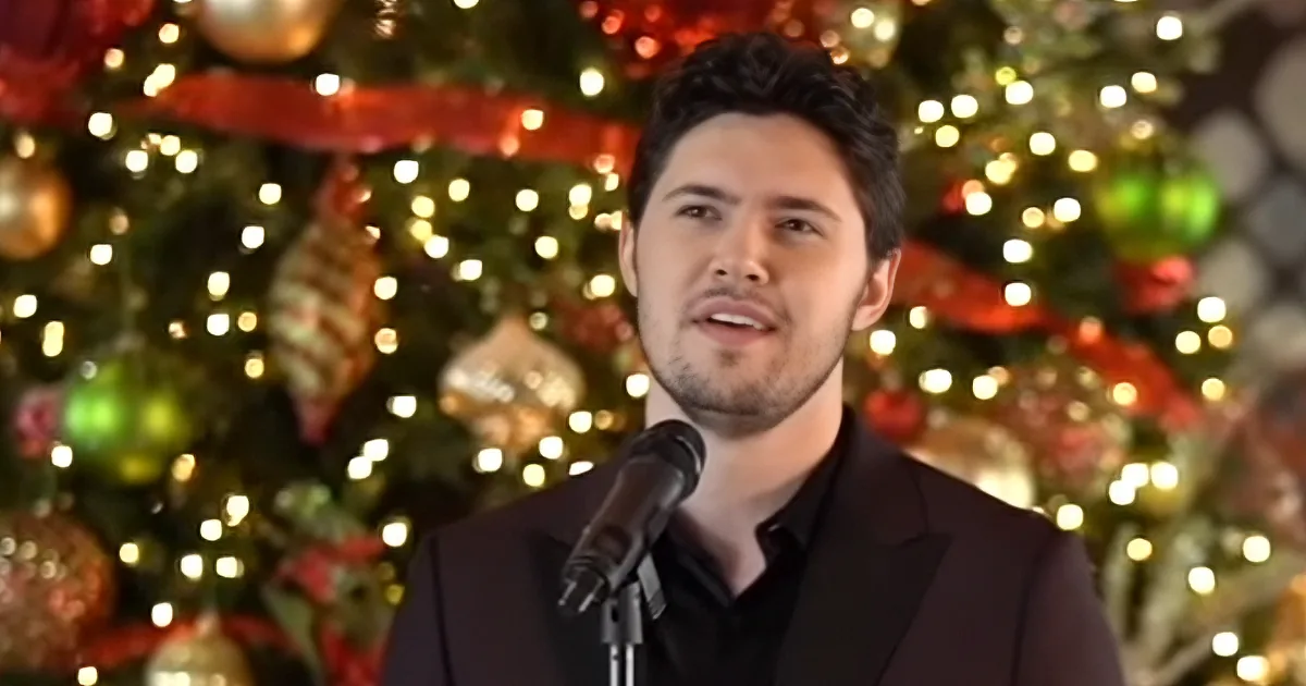 A man in a black suit sings into a microphone in front of a decorated Christmas tree.