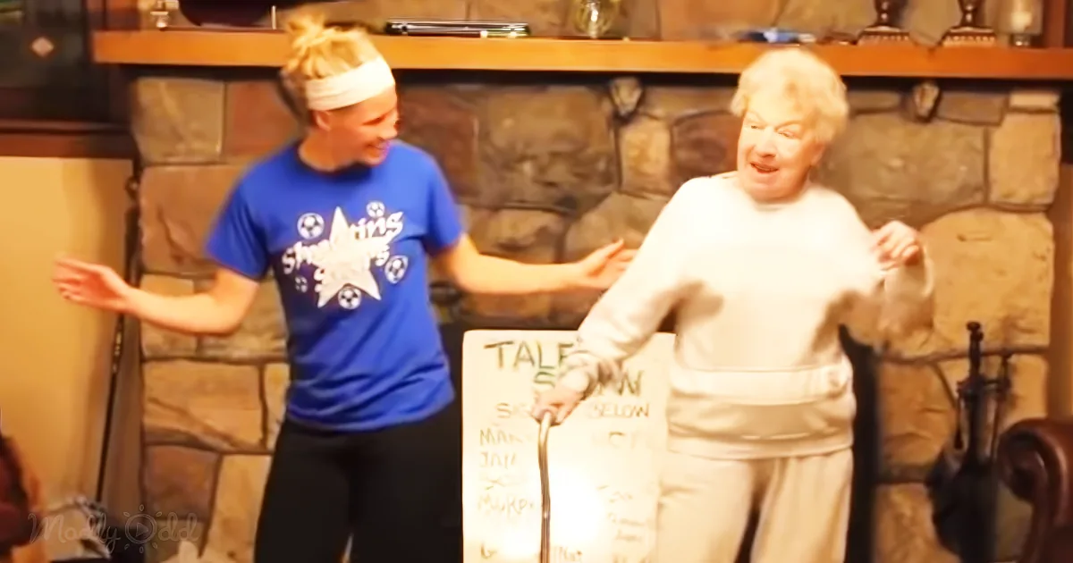 An elderly woman in white is dancing, holding a sign, while a younger woman in blue assists and smiles.