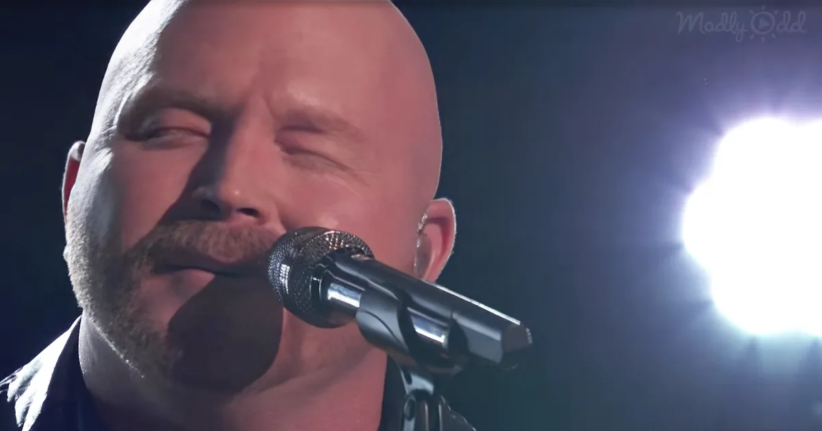 A bald man with a beard sings into a microphone under bright stage lights.