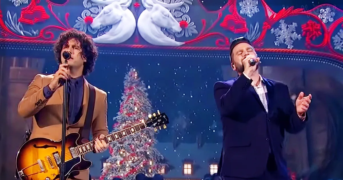 Two men perform on stage, one playing guitar, both singing into microphones, with a Christmas tree and festive backdrop.