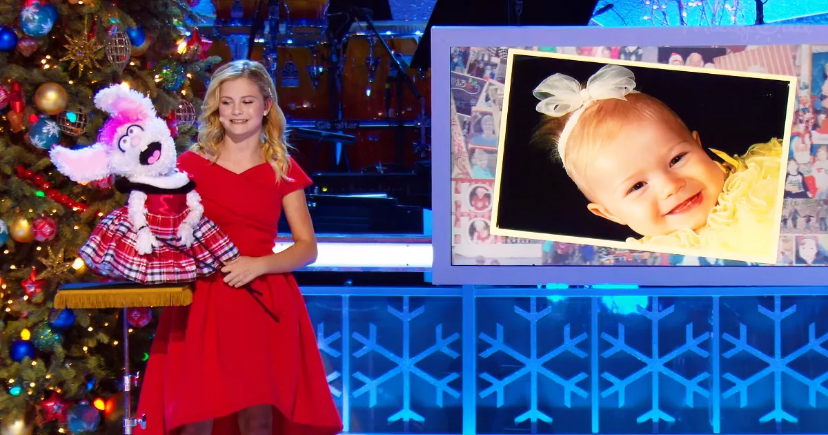 A young girl in a red dress holds a puppet, standing next to a Christmas tree and a picture of a baby.