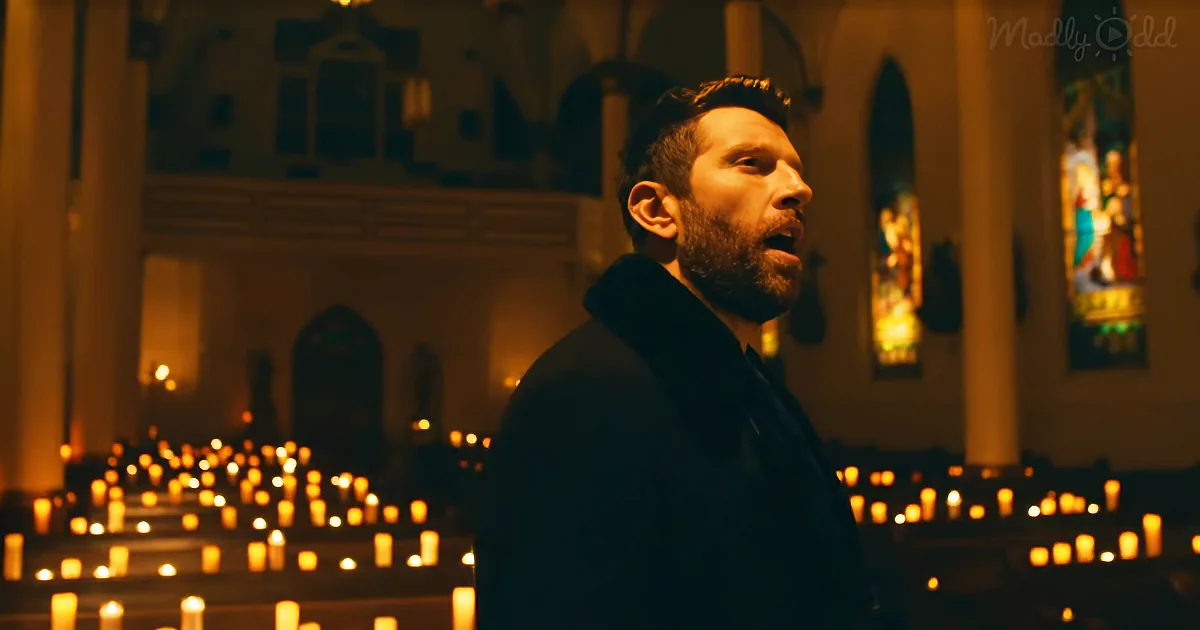 A man with a beard stands in a dimly lit church, surrounded by lit candles, looking to the right.