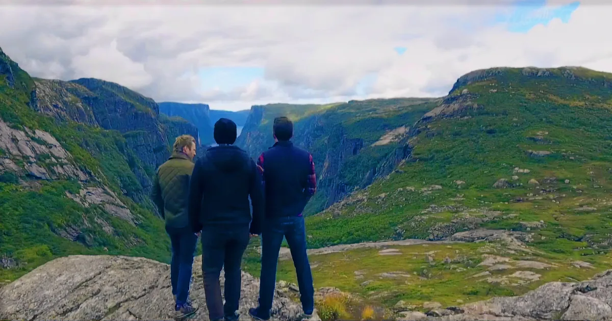 Three people stand on a rocky cliff overlooking a vast, green valley with steep cliffs and a cloudy sky.