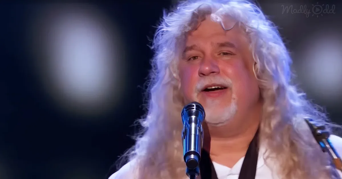 A man with long white hair and a beard sings into a microphone on stage.