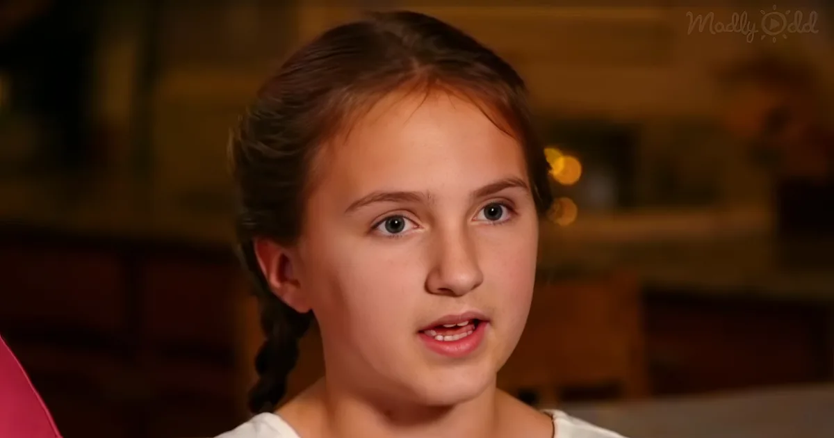 A young girl with braided hair and a white top is speaking, with a blurred background and a logo in the corner.