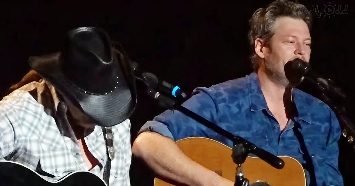 A man in a blue shirt plays guitar and sings into a microphone while another person in a cowboy hat listens.