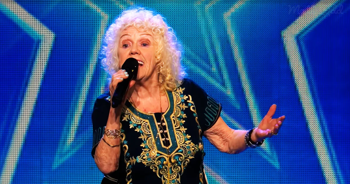 A woman with curly white hair sings into a microphone on stage, wearing a black and gold embroidered top.