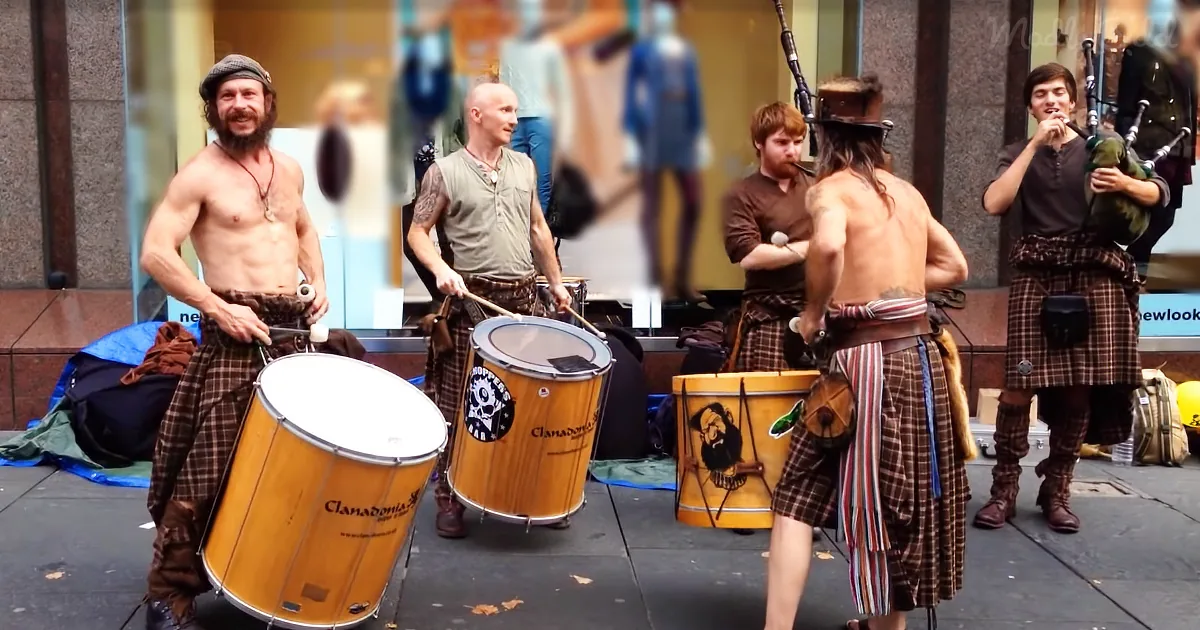 A man in a kilt plays a large drum with others in similar attire, creating a lively street performance.