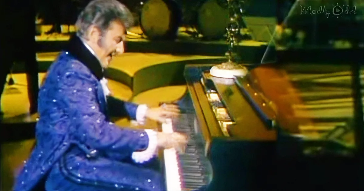 A man in a blue sequined suit plays a grand piano on a stage with yellow lighting.