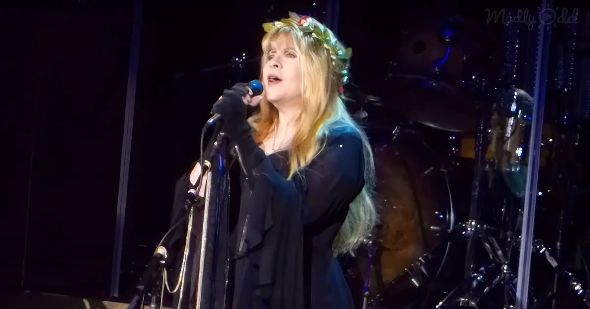 A woman in a black outfit and floral crown sings into a microphone on stage, with a drum set in the background.