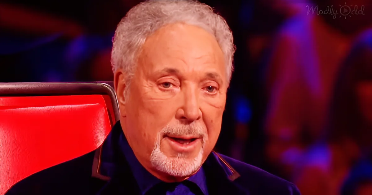 A man with white hair and a beard, wearing a dark suit, sits in a red chair, looking to the side.