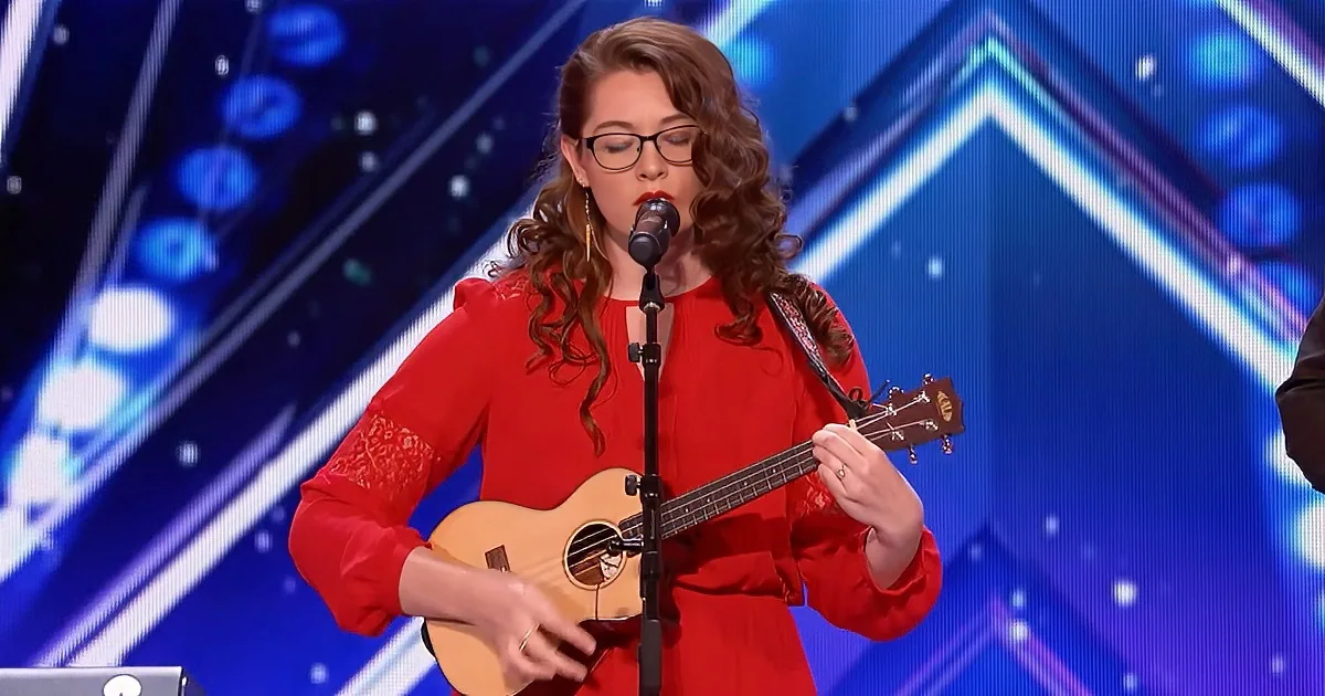 A woman in a red dress plays a ukulele and sings into a microphone on a stage with a blue geometric background.