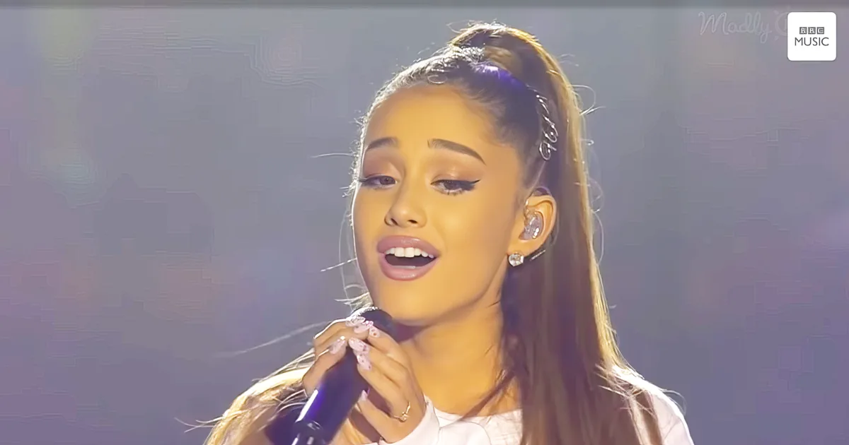 A woman with long hair in a ponytail, wearing a white top and earrings, sings into a microphone on stage.